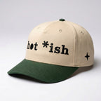 Hat with cream-colored crown and dark green colored bill that that says "hot *ish on front in stylized black embroidery, with black embroidered 4 point north star on left panel