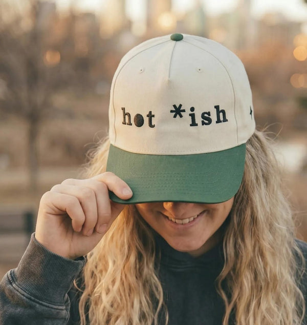 Model wearing Hat with cream-colored crown and dark green colored bill that that says "hot *ish on front in stylized black embroidery, with black embroidered 4 point north star on left panel, outdoor park environment in Minneapolis