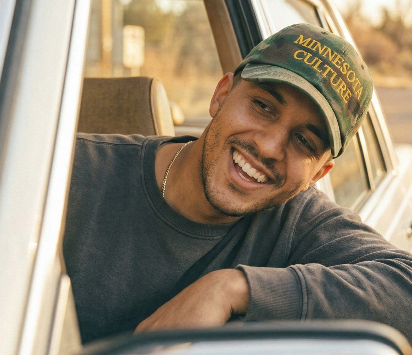 Model leaning out of driver side window of vehicle, wearing camo-colored MINNESOTA CULTURE hat with gold embroidered lettering