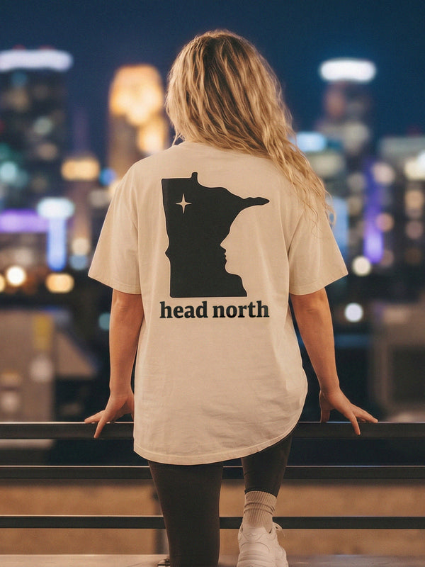 Model wearing beige oversized T-shirt with black minnesota logo, minneapolis skyline at night background