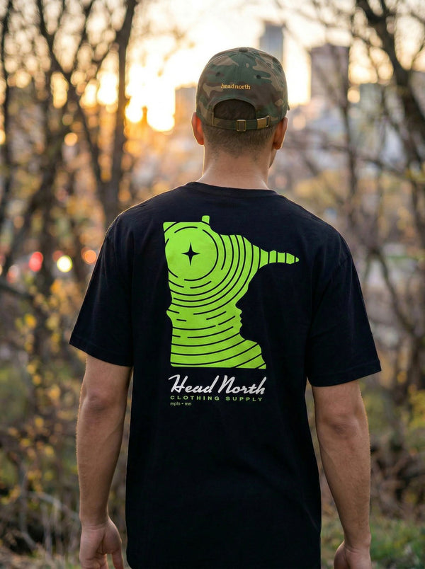 Model showing back of faded black T-shirt with green minnesota shaped design and Head North Clothing Supply text
