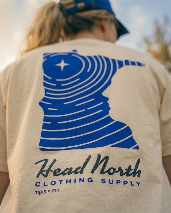 Model showing back of beige T-shirt with blue graphic of Minnesota and text Head North Clothing Supply